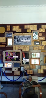 A wall display consisting of various framed artworks, photographs, and pieces of text is arranged in a collage-like manner. In front of the wall, an individual is sitting on a chair reading a newspaper. Some frames display images and others art or text in different languages. A set of blue ropes serves as a barrier in front of the wall.