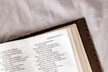 An open book displaying a page from the Bible, specifically the book of Zephaniah. The page is slightly tilted and the focus is on the text, which is in a serif font. The book has a leather cover and is placed on a soft, off-white surface.