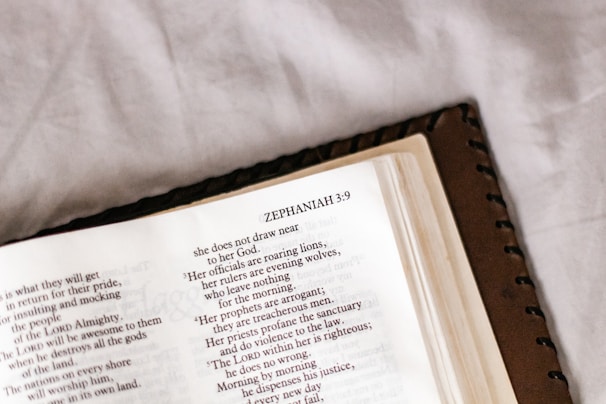 An open book displaying a page from the Bible, specifically the book of Zephaniah. The page is slightly tilted and the focus is on the text, which is in a serif font. The book has a leather cover and is placed on a soft, off-white surface.