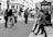 A busy city street scene with numerous people walking in different directions. The area is lined with buildings and shops, and a large poster with '15 AÑOS RODANDO EN MÁLAGA' in Spanish is prominently displayed. The image is in black and white, with shadows cast by the sunlight.