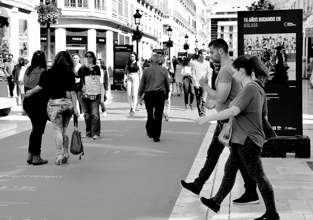 A busy city street scene with numerous people walking in different directions. The area is lined with buildings and shops, and a large poster with '15 A&Ntilde;OS RODANDO EN M&Aacute;LAGA' in Spanish is prominently displayed. The image is in black and white, with shadows cast by the sunlight.