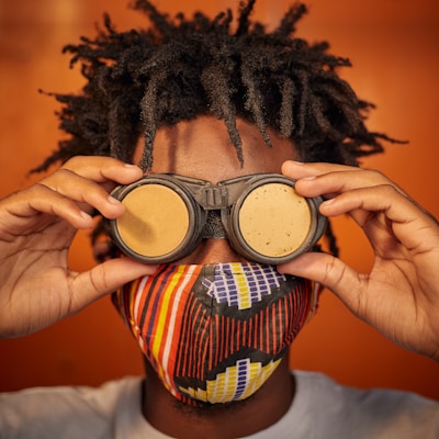 A person with curly hair is holding a pair of steampunk goggles over their eyes. They wear a colorful patterned face mask featuring stripes and geometric designs. The background is a warm, solid orange color, complementing the vibrant mask.