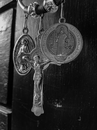 A black and white image of religious pendants hanging from a ring. The central pendant features a crucifix with a detailed depiction of Christ. Flanking the crucifix are two medallions with religious engravings and inscriptions surrounding figures.