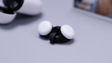 A pair of wireless earbuds with a sleek design, featuring white outer panels and black inner parts. The earbuds are placed on a smooth, light gray surface with a charging case seen in the background.