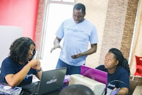 Three people are in a casual office setting. Two women are seated at a table working on laptops, while a man stands between them, engaging in conversation. They all appear focused and engaged in discussion. The environment looks collaborative.