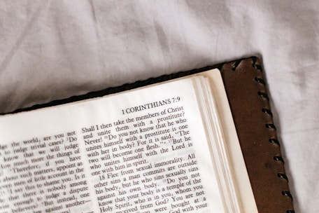 An open book featuring a page from the Bible with text from 1 Corinthians 7:9. The book has a brown leather cover and is resting on a light-colored surface.