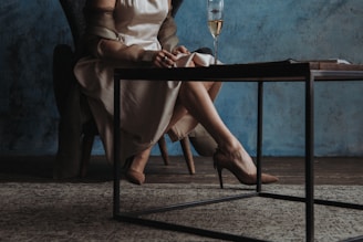 A woman dressed in a cream-colored satin dress and high heels is sitting on a chair with a blue backdrop. Her hands rest on her lap, and there is a glass of champagne on a low black metal table in front of her. The room has a rustic wooden floor and a patterned rug and an open book is also on the table.