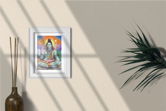 A framed artwork featuring a colorful depiction of a deity is mounted on a beige wall. Natural light casts soft shadows across the surface. To the left, a narrow vase contains a bundle of tall, thin sticks. To the right, a green leafy plant adds a touch of nature to the scene.