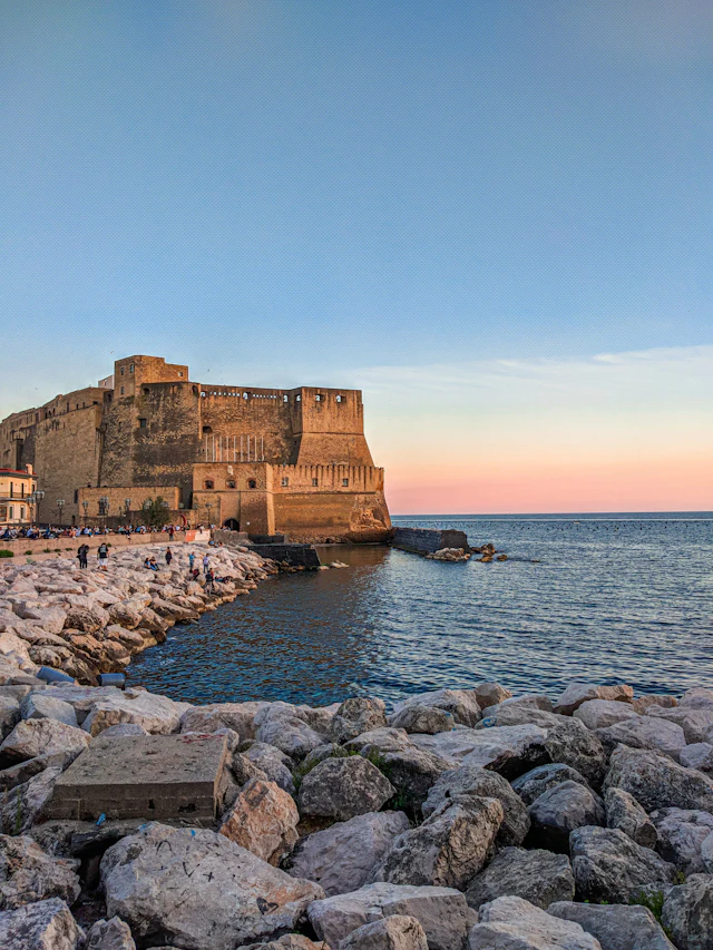 Castel dell'Ovo on its rocky seafront promontory at sunset, with people strolling the Lungomare below.