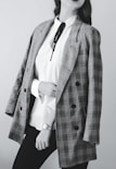 A person dressed in a stylish outfit featuring a plaid blazer draped over the shoulders. Underneath, a white blouse with a black tie detail is paired with dark pants. The person wears a wristwatch.