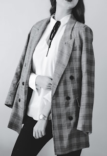 A person dressed in a stylish outfit featuring a plaid blazer draped over the shoulders. Underneath, a white blouse with a black tie detail is paired with dark pants. The person wears a wristwatch.