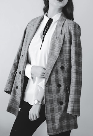 A person dressed in a stylish outfit featuring a plaid blazer draped over the shoulders. Underneath, a white blouse with a black tie detail is paired with dark pants. The person wears a wristwatch.
