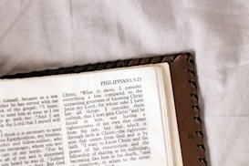 An open page of a book with the passage from Philippians 3:21. The text is clear and printed in black on white paper. The page is resting on a surface that appears to be a brown leather cover and a white fabric background.