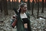 A person stands in a dimly lit forest, holding a rifle over their shoulder. They wear a dark top and a green jacket, with their hair tied up. The forest floor is covered in dry leaves, and the background is filled with bare trees and a hint of an orange sunset peeking through.