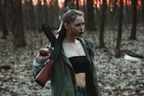 A person stands in a dimly lit forest, holding a rifle over their shoulder. They wear a dark top and a green jacket, with their hair tied up. The forest floor is covered in dry leaves, and the background is filled with bare trees and a hint of an orange sunset peeking through.