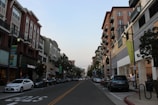 A city street lined with modern multi-story buildings featuring a mix of retail stores with signs such as Fossil and The Body Shop. Cars are parked along both sides of the street, and a few people can be seen walking on the sidewalks. The sky is clear, with the evening light creating a calm atmosphere.
