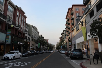 A city street lined with modern multi-story buildings featuring a mix of retail stores with signs such as Fossil and The Body Shop. Cars are parked along both sides of the street, and a few people can be seen walking on the sidewalks. The sky is clear, with the evening light creating a calm atmosphere.