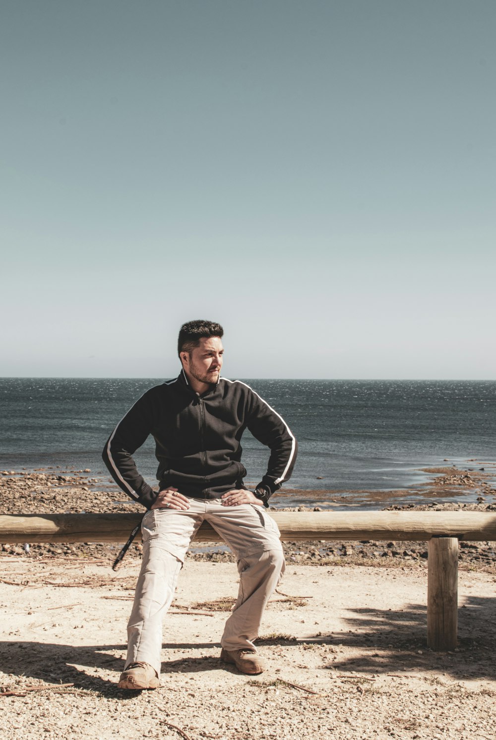 man in black jacket and white pants sitting on brown wooden fence near sea during daytime