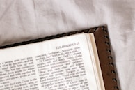 An open page of a book, likely a Bible, featuring the heading 'COLOSSIANS 3:25' with text in a serif font. The book is placed on a white, textured surface and partially covered with a brown leather cover that has a stitched border.