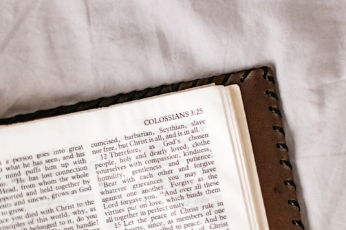 An open page of a book, likely a Bible, featuring the heading 'COLOSSIANS 3:25' with text in a serif font. The book is placed on a white, textured surface and partially covered with a brown leather cover that has a stitched border.