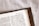 An open page of a book, likely a Bible, featuring the heading 'COLOSSIANS 3:25' with text in a serif font. The book is placed on a white, textured surface and partially covered with a brown leather cover that has a stitched border.