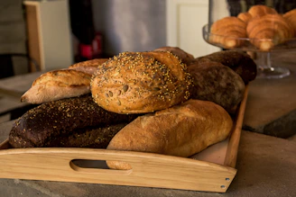A wooden tray filled with an assortment of freshly baked bread, featuring different textures and toppings such as seeds. The bread includes a variety of types, including a dark loaf and several light, crusty loaves. In the background, a glass cake stand holds a selection of croissants.