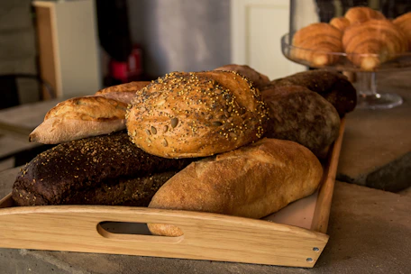 A wooden tray filled with an assortment of freshly baked bread, featuring different textures and toppings such as seeds. The bread includes a variety of types, including a dark loaf and several light, crusty loaves. In the background, a glass cake stand holds a selection of croissants.