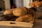 A wooden tray filled with an assortment of freshly baked bread, featuring different textures and toppings such as seeds. The bread includes a variety of types, including a dark loaf and several light, crusty loaves. In the background, a glass cake stand holds a selection of croissants.