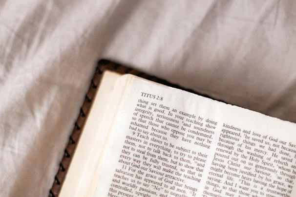 An open book resting on a textured fabric surface, featuring text from a religious passage with a prominent title 'TITUS 2:8'. The page is slightly creased and the lighting is soft, casting gentle shadows.