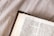 An open book resting on a textured fabric surface, featuring text from a religious passage with a prominent title 'TITUS 2:8'. The page is slightly creased and the lighting is soft, casting gentle shadows.
