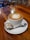 A cup of coffee with intricate latte art served on a white saucer, accompanied by a small metallic spoon and a packet of sugar labeled 'mawana', placed on a polished wooden table.