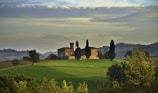 A picturesque landscape featuring a rustic villa on a hill surrounded by grass fields and tall cypress trees. The background showcases a range of rolling hills and a cloudy sky, creating a serene and timeless rural scene.