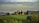 A picturesque landscape featuring a rustic villa on a hill surrounded by grass fields and tall cypress trees. The background showcases a range of rolling hills and a cloudy sky, creating a serene and timeless rural scene.
