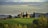 A picturesque landscape featuring a rustic villa on a hill surrounded by grass fields and tall cypress trees. The background showcases a range of rolling hills and a cloudy sky, creating a serene and timeless rural scene.