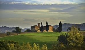 A picturesque landscape featuring a rustic villa on a hill surrounded by grass fields and tall cypress trees. The background showcases a range of rolling hills and a cloudy sky, creating a serene and timeless rural scene.