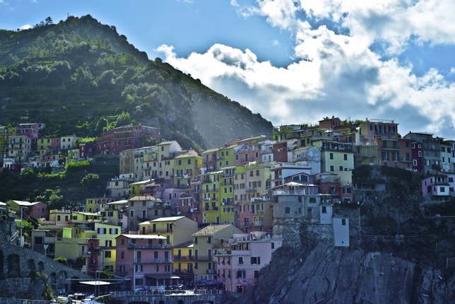 A coastal village with colorful buildings clinging to a rocky hillside. The vibrant homes are painted in various shades of pink, yellow, orange, and white, creating a picturesque contrast against the lush green mountains and the bright blue sky filled with fluffy clouds.