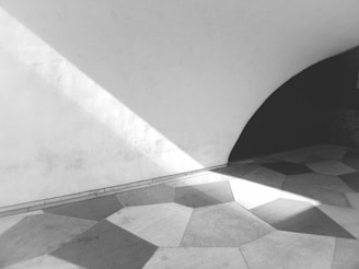 Geometric patterns of shadows and light fall across a tiled floor. The tiles are irregularly shaped, and the design is minimalist with a stark contrast between light and dark. The curved wall in the background adds an element of architectural interest.