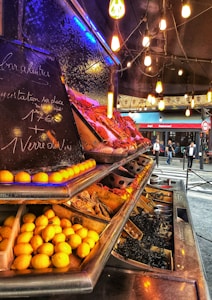 An outdoor seafood market display with a variety of fresh fruits and seafood on ice. The counter is lined with lemons and various other foods. The setting is warmly lit with hanging bulbs, creating an inviting ambiance. Chalkboard signs provide information about goods for sale.