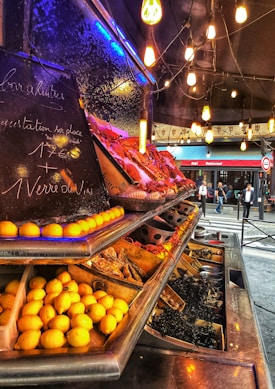 An outdoor seafood market display with a variety of fresh fruits and seafood on ice. The counter is lined with lemons and various other foods. The setting is warmly lit with hanging bulbs, creating an inviting ambiance. Chalkboard signs provide information about goods for sale.