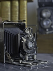 An old-fashioned accordion-style camera is prominently displayed, featuring detailed metal and leather craftsmanship. In the background, there is a stack of books and another vintage camera with dual lenses.