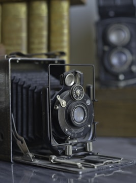 An old-fashioned accordion-style camera is prominently displayed, featuring detailed metal and leather craftsmanship. In the background, there is a stack of books and another vintage camera with dual lenses.