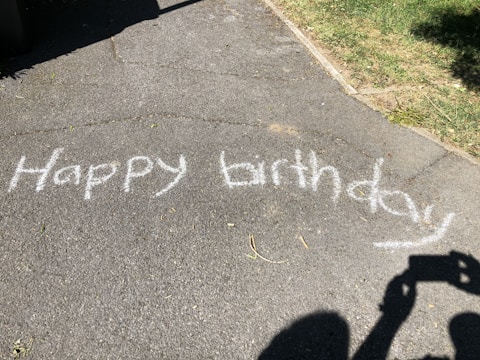 White chalk letters spell 'Happy birthday' on a paved surface, with grass visible on the right side. A shadow of a person taking a photo is visible in the bottom right corner.
