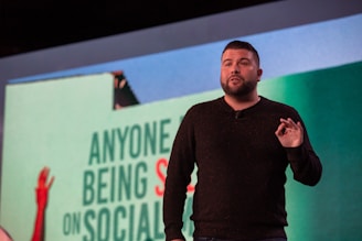 A person is speaking on a stage, likely giving a presentation. They are gesturing with one hand and are possibly emphasizing a point. Behind them is a large screen displaying partial text that appears to discuss social media.