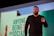 A person is speaking on a stage, likely giving a presentation. They are gesturing with one hand and are possibly emphasizing a point. Behind them is a large screen displaying partial text that appears to discuss social media.
