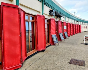 A row of red wooden shutters line the exterior of a building, with large glass windows in between. The shutters are open and neatly aligned. Above the windows, there is a green sign featuring the words 'Littleton Wine Merchants.' Two sandwich board signs are placed on the pavement advertising drinks, such as beers and wines.