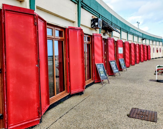 A row of red wooden shutters line the exterior of a building, with large glass windows in between. The shutters are open and neatly aligned. Above the windows, there is a green sign featuring the words 'Littleton Wine Merchants.' Two sandwich board signs are placed on the pavement advertising drinks, such as beers and wines.
