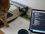 A ginger cat is attentively looking at a sketch of a shirtless man on a drawing pad. The scene is set on a desk where a vintage typewriter, a laptop displaying code, and various stationery items are present. The blue cup in the foreground indicates a relaxed or creative work environment.