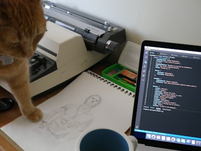 A ginger cat is attentively looking at a sketch of a shirtless man on a drawing pad. The scene is set on a desk where a vintage typewriter, a laptop displaying code, and various stationery items are present. The blue cup in the foreground indicates a relaxed or creative work environment.