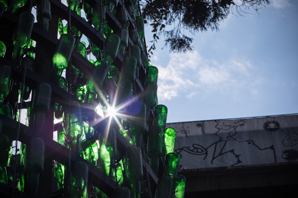 Green glass bottles are arranged in a lattice pattern, with sunlight streaming through them, creating a starburst effect. The background features a partly cloudy sky and graffiti art on a wall.