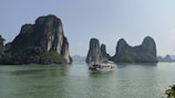 A scenic view of towering limestone islands covered with lush greenery rises from calm, turquoise waters. A traditional boat with a flag and several passengers sails across the water, creating a serene landscape.